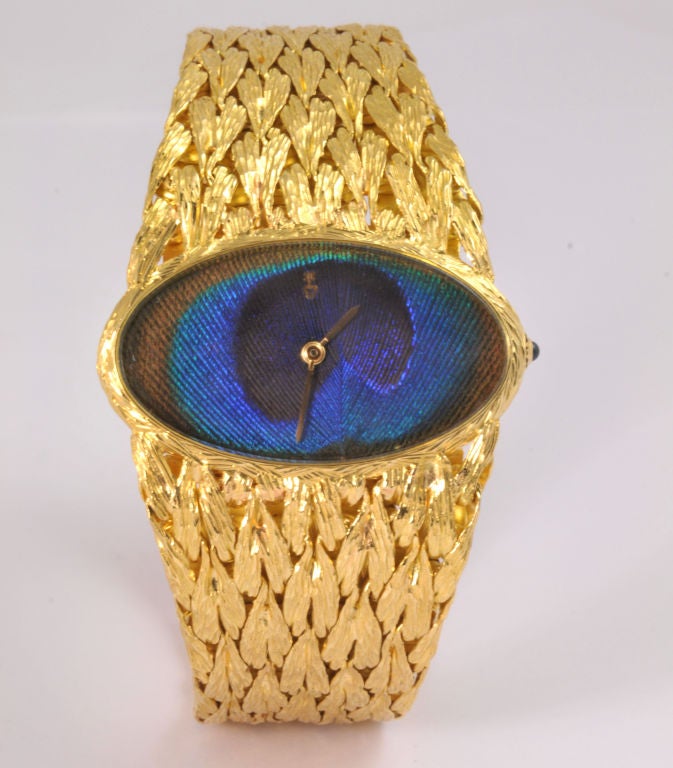 An 18K yellow gold watch with a blue enamel peacock feather in the watch face and a blue sapphire.