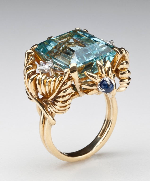 An 18 karat gold ring, circa 1960-70, by Jean Schlumberger for Tiffany.  The 14 carat aquamarine measures 16.3mm by 13.5mm by 9.4mm and is flanked by a foliate design set with two full cut diamonds, a cabochon sapphire and a cabochon emerald.  