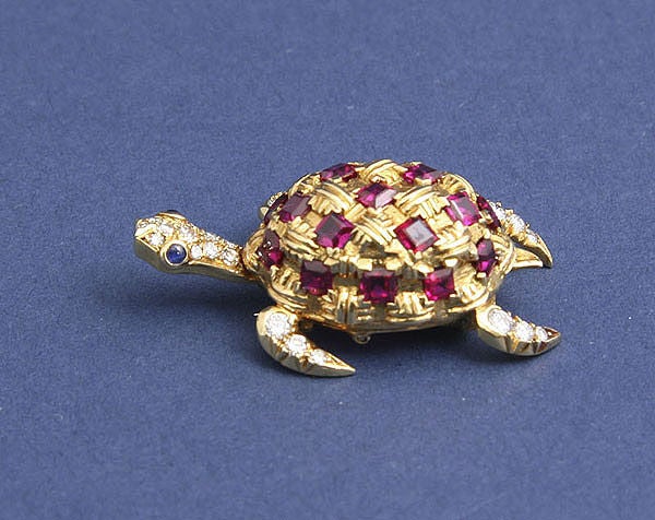An 18 karat gold clip brooch of a turtle with rubies, diamonds and sapphires.  The turtle's head moves to the right and the left; the back is studded with approximately one carat of rubies, the neck and head is covered with tiny diamonds; the eyes