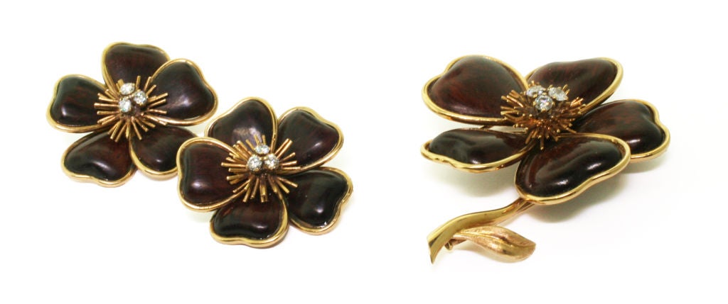 Comprising; a brooch and pair of ear clips, the brooch of <br />
flower head design, centring on a trio of brilliant-cut diamonds, to <br />
carved wooden petals and polished gold stem, together with a pair of <br />
ear clips en suite. Signed