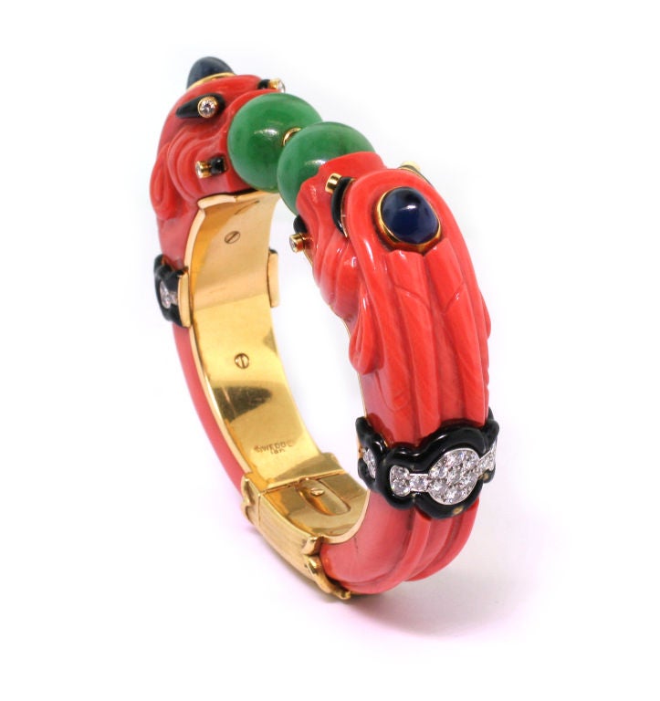 The carved coral bangle embellished with brilliant-cut diamonds within black enamel surrounds, to two chimera heads each accented with a cabochon sapphire, centring on two jade beads, circa 1965, signed Webb, stamped 18k for 18ct gold.