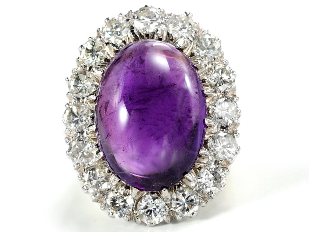 A divine hue of amethyst, a true royal purple awaits the viewer of this more than 11 carat gemstone. Cut en cabochon (flat bottom and smooth rounded top), the oval stone is mesmerizing with its surround of about 2 carats of luscious transitional cut