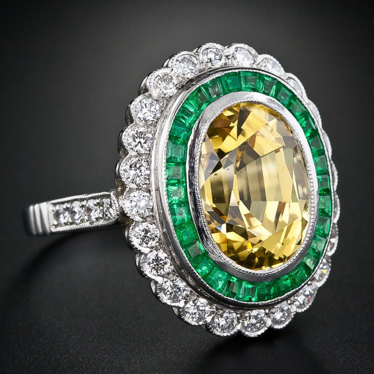 This big, bold and colorful ring centers on an 9.00 carat oval-shaped rich golden-yellow sapphire, surrounded by a double bezel of bright green calibre-cut emeralds and round brilliant-cut diamonds with a scalloped border. This handmade platinum