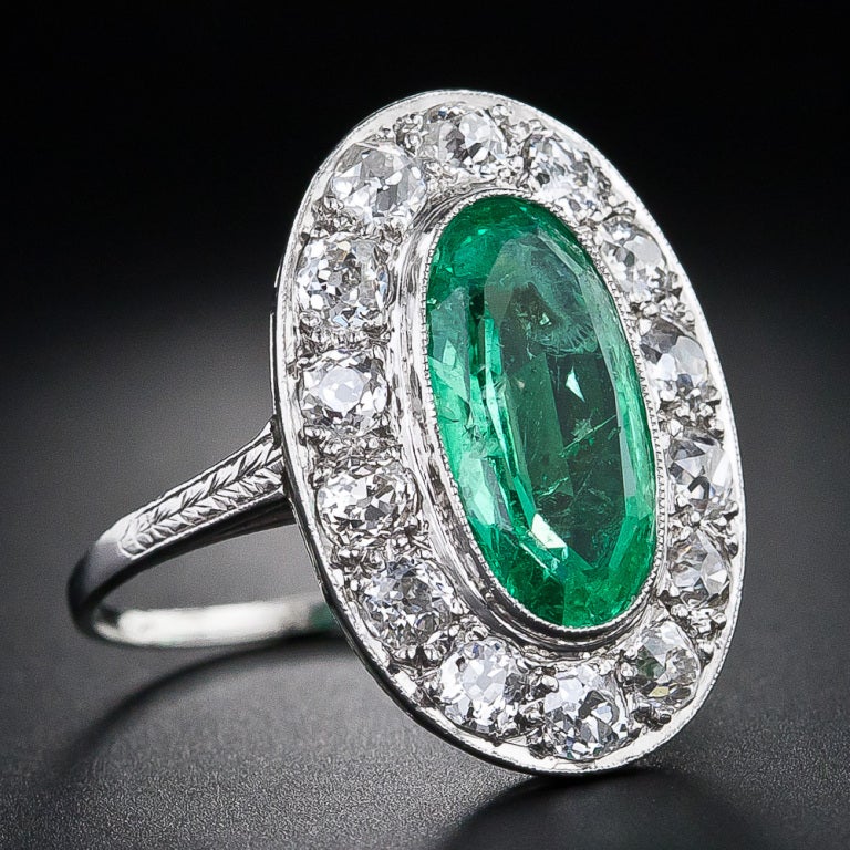 This glorious - 1 and 3/8 inch long - original Art Deco ring, crafted in platinum circa 1920s, glistens front and center with a bright and beautiful, crystalline green, elongated oval emerald, weighing 2 carats. The emerald is embraced all around