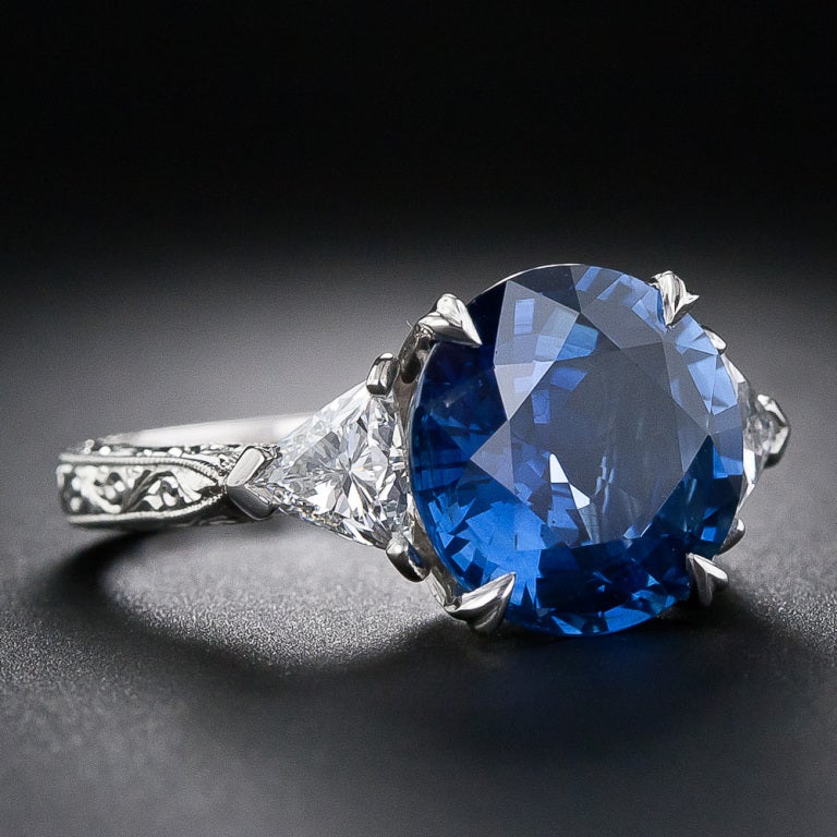 A sizable 6.62 carat round cut sapphire is the star of this vintage style platinum ring. The sapphire is embraced simply by a triangle cut diamond on either side. The ring is finished with a hand engraved scroll motif. Lovely.

Inventory No.