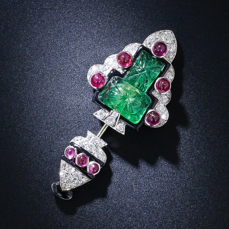A consummate, colorful and exotic jabot pin from the high Art Deco period - circa 1925. This magnificent Cartier-esque jewel, evocative of a festive Christmas tree, is composed of a pair of carved Moghul emeralds outlined in dramatic black enamel.
