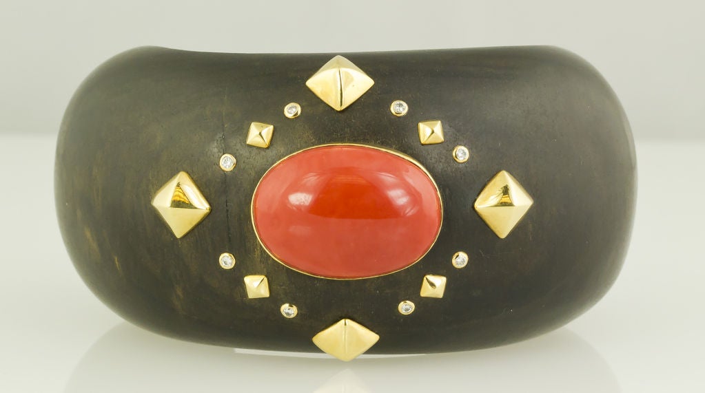Chic and interesting cuff bracelet by Trianon. It features a beautiful cabochon coral center stone, approx. 15mm x 12mm accented by bezel set diamonds and 18K gold pyramid shaped beads. Made in ebony wood. <br />
Hallmarks: Trianon, 750.