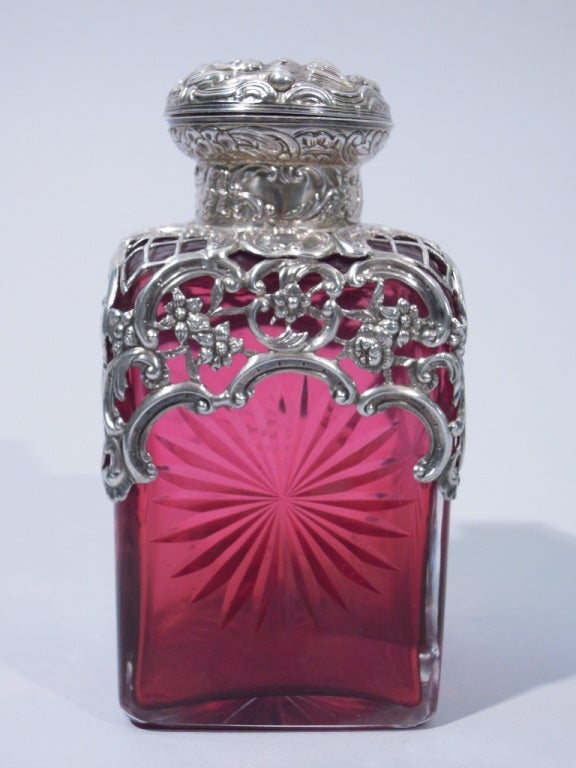 This unusual perfume bottle was made in London Circa 1894. It has a square shape and is four sided. The body of the bottle is done with very rare red color glass which is cut on all four sides and the bottom in starburst patterns. The top section of
