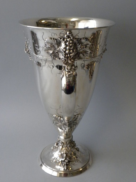 This monumental Antique American Sterling Silver Two Handled Trophy Cup is decorated with an ornate repousse grape vine motif consisting of large figural grapes, vines and leaves as well as vine twigs flowing from the branches.  The design is