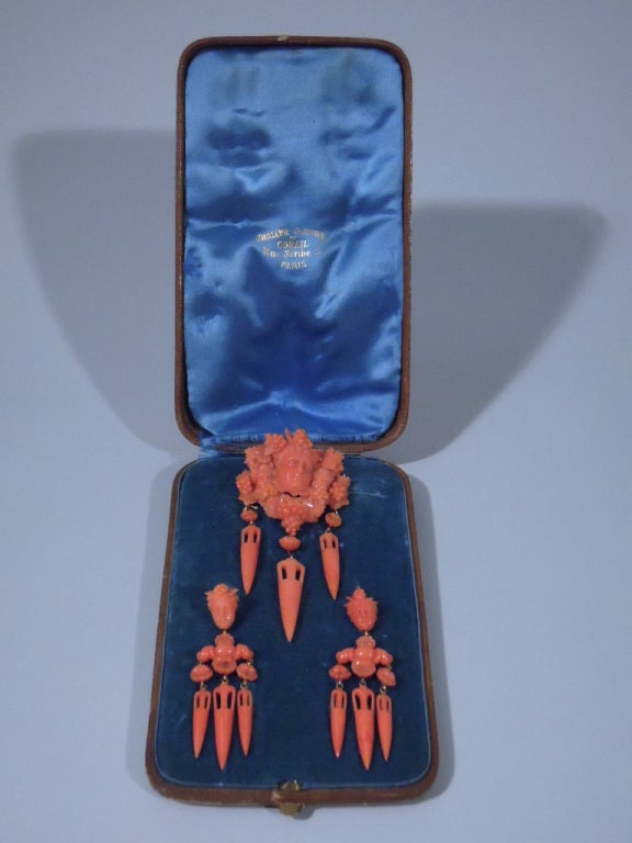 A suite of coral earrings and brooch with carved Classical masks on 18 kt gold mounts. The brooch has a male mask surrounded by grape bunches and 3 pierced dagger pendants. The earrings have a mask with pendant beads, shells, and 3 pierced daggers.
