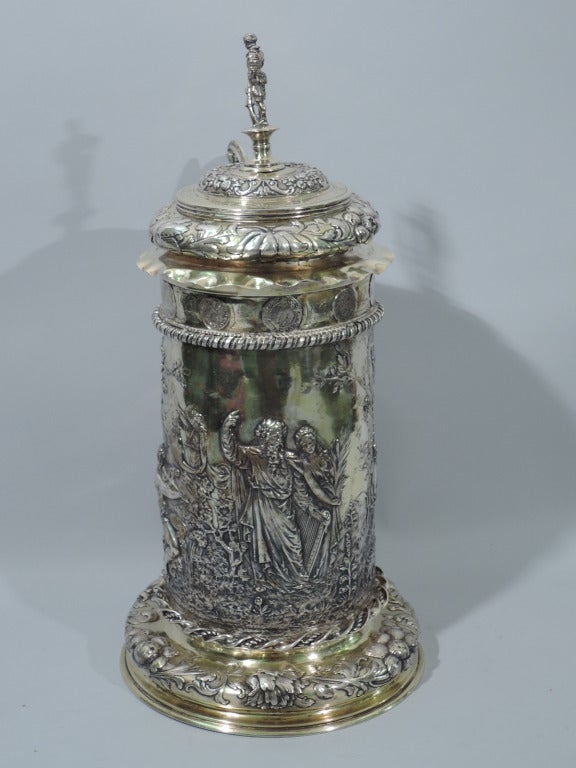 German silver gilt tankard with Classical frieze, ca. 1880. Tapering cylinder, scroll handle, double-dome hinged cover with piecrust rim, and bun foot. Youths and nymphs dance, lounge, and play music in the presence of a serene elder sage. Floral