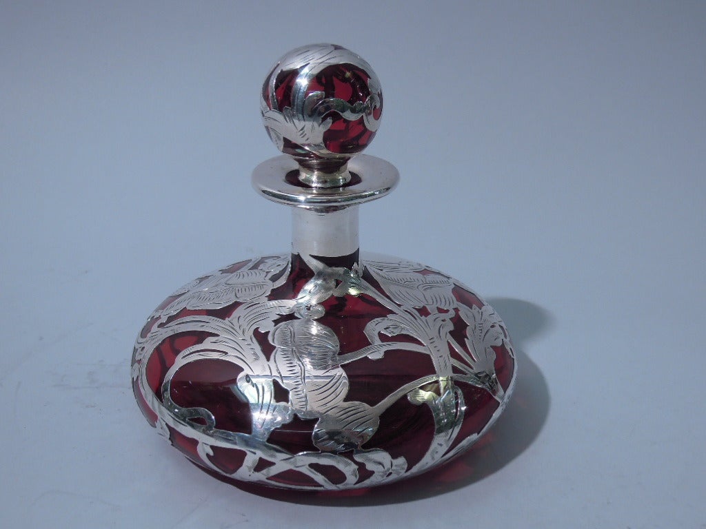 American perfume bottle in ruby glass with silver overlay, c1900. Bottle has squat and curved bowl, cylindrical neck, foot ring and everted rim. Ball finial with short and tapering plug in ruby shading to clear. Asymmetrical overlay with flowers and