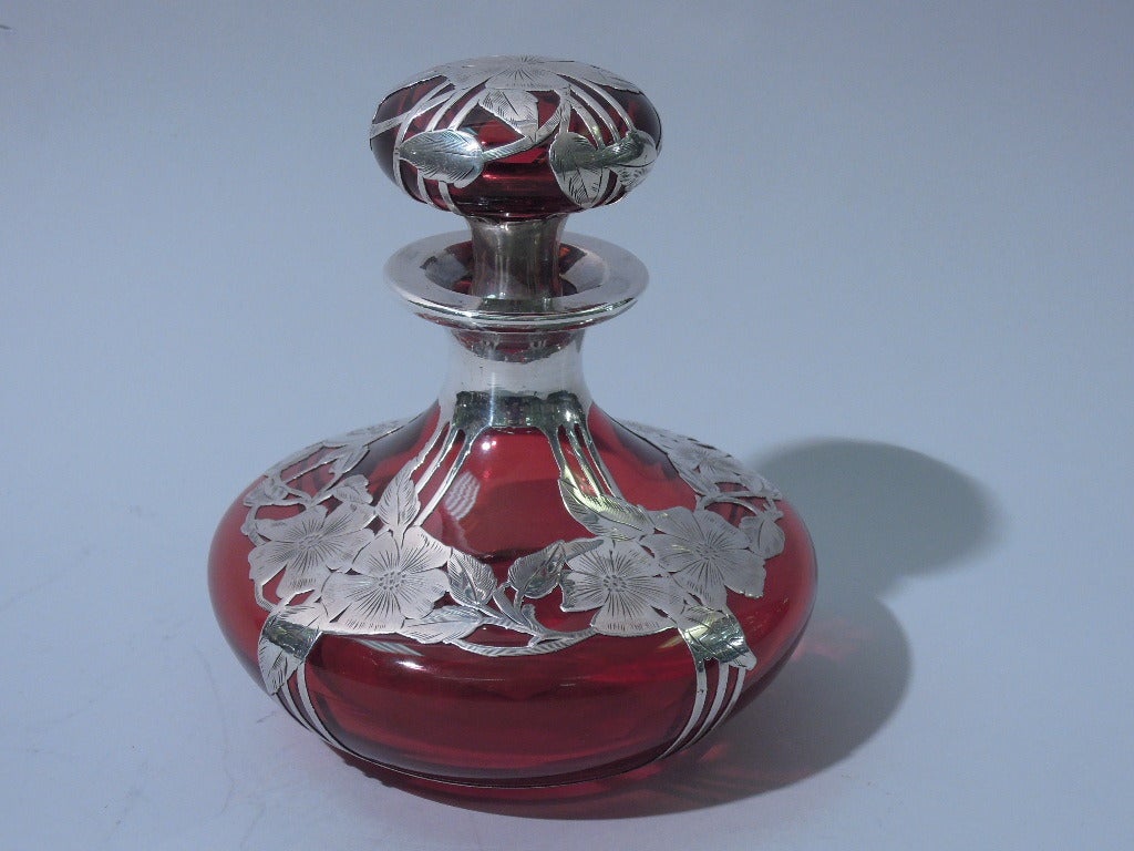 Large perfume bottle in cranberry glass with 999 silver overlay. Made by Alvin in Providence, c1900. Bottle has squat and curved bowl, short neck and everted rim. Stopper is squat and curved with short and tapering plug in cranberry shading to