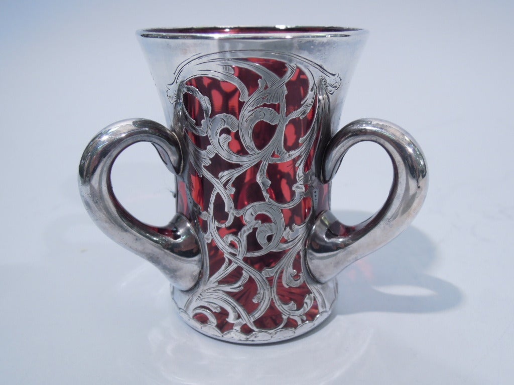 American three-handled loving cup in cranberry glass with silver overlay, c1900. Concave cylinder with three scrolled handles. Asymmetrical overlay with scrolls and heraldic cartouche (blank for engraving). Excellent condition.
 
Diminutive