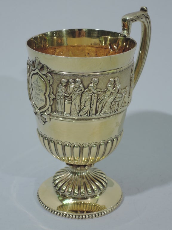 Victorian sterling silver gilt mug. Made by George Angell in London in 1874. Bowl has Classical frieze with harp-playing, courting, and reclining figures. Bowl bottom is lobed as is domed and beaded foot. Scrolled bracket handle is reeded with