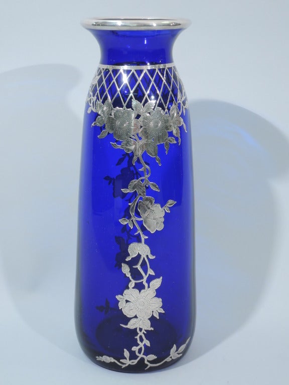 Large cobalt glass vase with silver overlay, ca. 1910. Straight and upward tapering sides and flared rim. Top encircled with silver trellis supporting a dangling garland on front and back. Rim has silver collar. A controlled and vertical design that