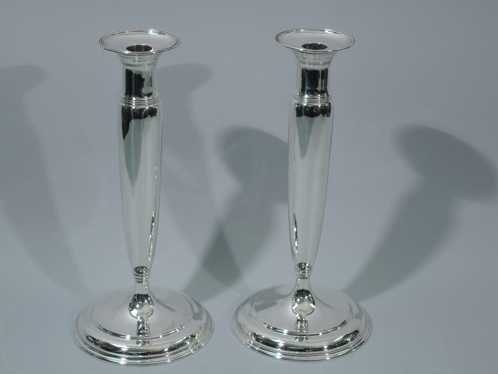 Pair of large sterling silver candlesticks. Made by Tiffany in New York, ca. 1907. Tapering support mounted to stepped dome foot with dentil border. Support surmounted by cylindrical neck with stepped and reeded flared rim. Bobeche has beaded rim.