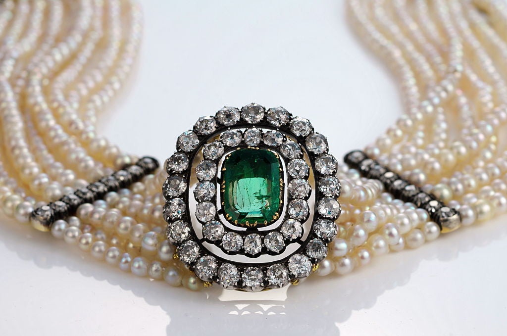 A magnificent diamond, emerald and natural pearl dog collar, the centerpiece set with a Colombian emerald, mounted in gold and surrounded by two rows of old mine cut diamonds, attached to a 9-strand natural pearl necklace, interspersed with 2 old