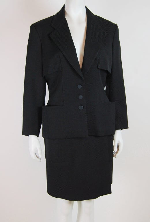 Gorgeous early 90's minimal style suit set in diagonal ribbed wool. Unlined wrap skirt fastens with 2 buttons at the waist and has one pocket. Fitted jacket with wide lapel and capelet back. Matte black plastic buttons on closure, at pocket back