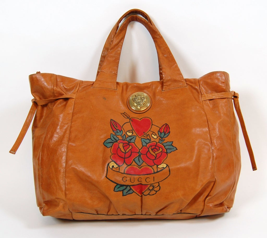 An impressively large leather tote from Gucci featuring a classic tattoo-style design with hearts and roses. Oversized gold-tone metal Gucci crest medallion. Leather ties at each side. Top zip closure. Lined in linen with one interior zipper pocket.