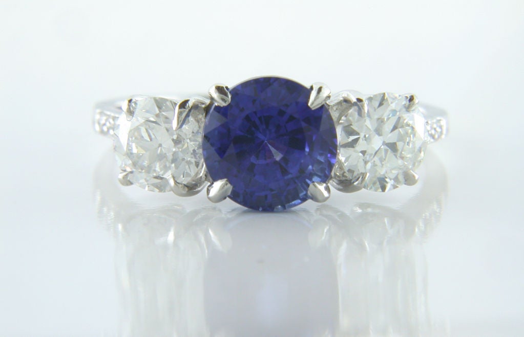 This lovely platinum three stone ring is a combination of both classic elegance and vintage style. The ring features a single round brilliant cut vivid blue sapphire weighing 2.74 carats. The center stone is flanked by two old european cut diamonds