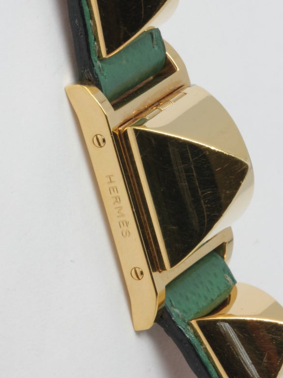 Hermes lady's gold-filled Medor wristwatch with concealed dial, 23mm width, cover lifts to reveal watch dial, with two similarly shaped decorative elements secured to green Hermes strap with gold-filled Hermes buckle. Quartz movement. Preowned in