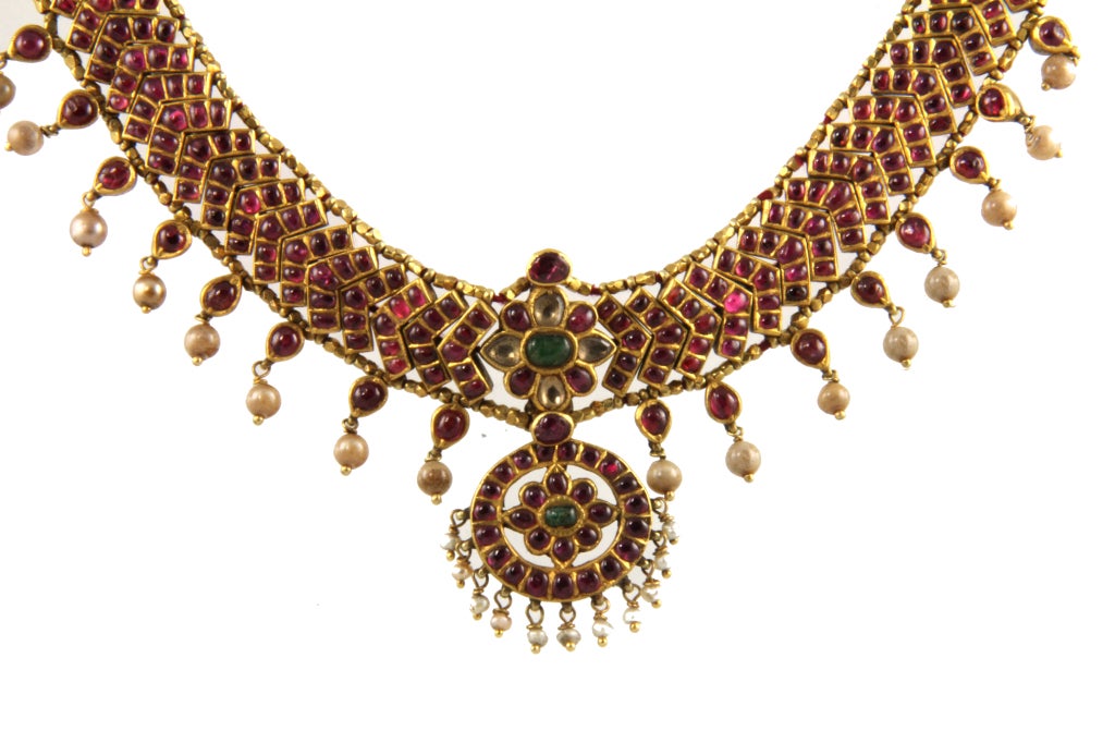 This 22 k Kundan Necklace is comprised of many Rubies set into the design. In the center there is also Emerald and Diamond. There are small  drop pearls around the bottom edge of the necklace. The necklace is adjustable and can be worn as a choker