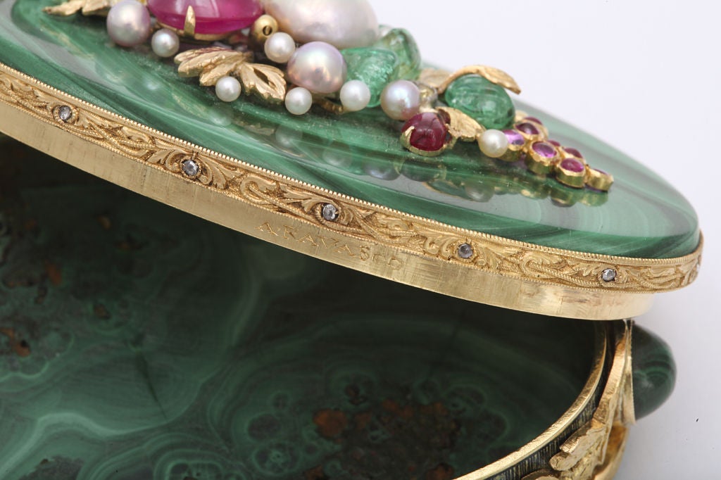 An ornate oval malachite box decorated with a cluster of cabochon emeralds, rubies and pearls on the lid and carved 18 karat gold around the outer circumference enhanced by rose cut diamonds and four cabochon malachite ports.