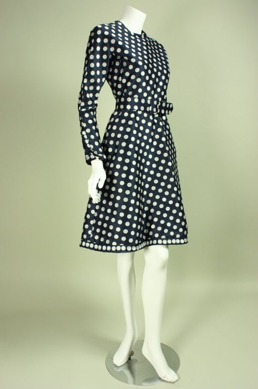 Vintage cocktail dress from Geoffrey Beene dates to the 1960's.  It is made of navy silk with an allover white polka-dotted print.  Fitted bodice has round neck and long, tapered sleeves with pleated snap cuffs.  Flared a-line skirt has same pleated
