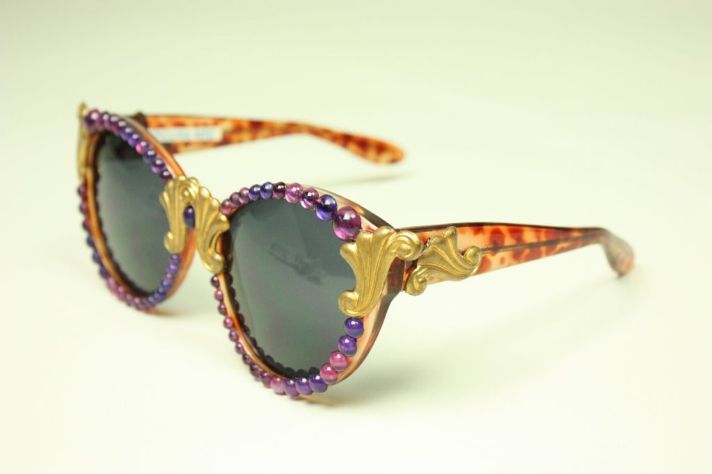 Contemporary sunglasses from Mercura NYC feature violet cabochons with scrolling gold-toned metal accents.  Tortoiseshell plastic frames.  Gray lenses.