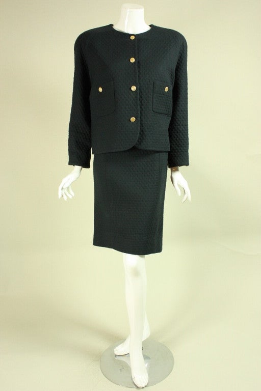 Vintage suit from Chanel dates to the 1980's and is made of black quilted cotton.  Boxy jacket has round neck, patch pockets, long sleeves with buttoned cuffs, and gold-toned buttons with the interlocking C's logo.  Straight skirt has center back