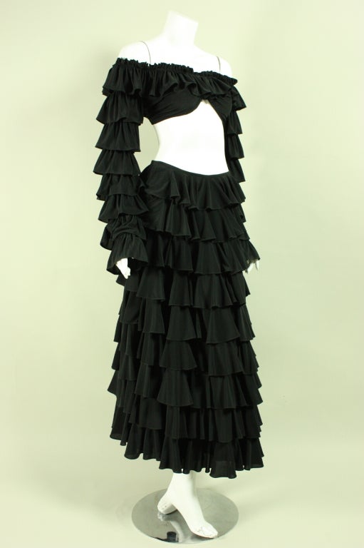Vintage ensemble from Norma Kamali's OMO line dates to the early 1980's.  It is made of tierred black ruffled fabric.  The off the shoulder crop top has a sweetheart neckline, elasticized openings, and long sleeves.  The ankle-length skirt has a