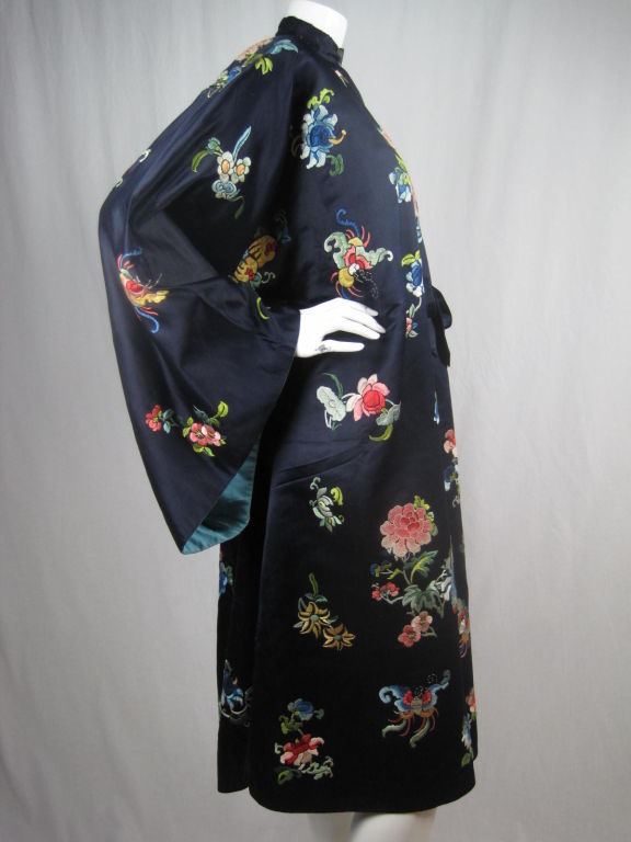 Chinese robe circa 1930-1950 likely made as an export for the Western market.  Navy satin has allover brightly colored, hand embroidery in a mainly floral motif. Single front tie at waist.  Mock neck fastens with two etched, brass-toned, and