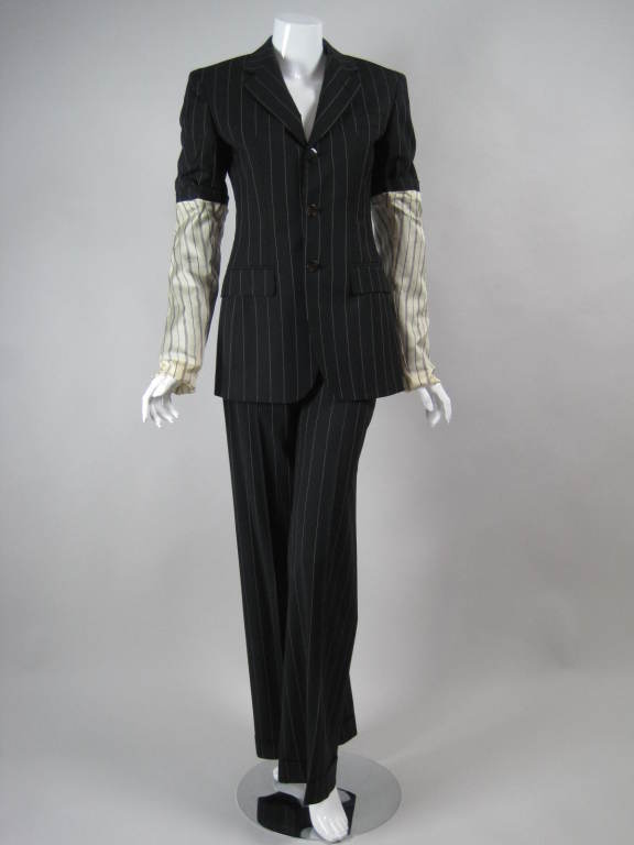 Two-piece trouser suit from Jean Paul Gaultier circa 1990's is made out of black pinstriped wool gabardine.  Single-breasted blazer has exaggerated turned back sleeve that exposes beige lining past the elbows.  Trousers have zip closure, flat front,