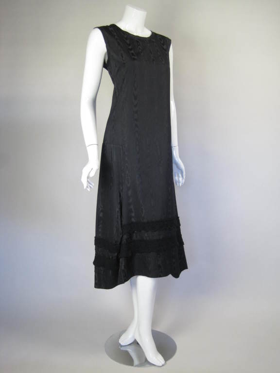 Minimalist sheath dress from Comme des Garcons is made out of black silk moiré.  It has a round neckline, is sleeveless, and below knee-length.  Side panels are inserted at hip level and flare outwards towards the front and back centers of the