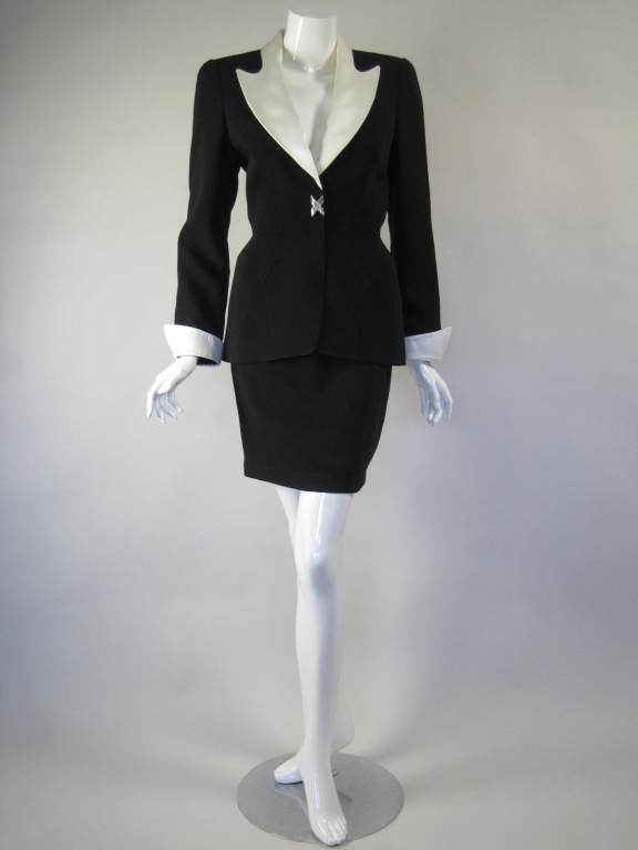Two-piece skirt suit from Thierry Mugler is made of of black wool faille and has a white satin collar and cuffs.  Single-breasted blazer has covered button closure and a rhinestone 