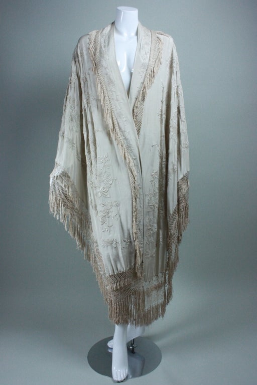 Circa 1920's cape is made from an embroidered Chinese export piano shawl.  It is made out of cream-colored silk dupioni with cream hand-embroidery in a floral motif.   Shawl lapel with narrow fringed edges.  Hem has fringed edges with macrame. 