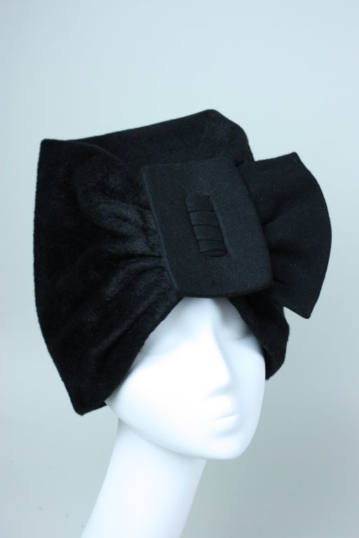 1950's through 1960's Surrealist-inspired hat from Lilly Dache is made out of velvety-soft black felt. It has turban-like styling with a large felt-covered buckle at the center front through which the fabric is pulled. 