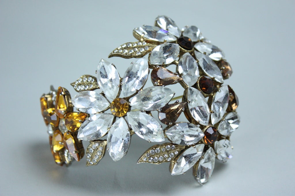 Ornate brooch from Christian Lacroix is made out of various shaped, faceted rhinestones in clear and amber that are set in gold-toned metal.  Brooch is arced and depicts floral sprays.  Leaves are set with rhinestones arranged in a pavé pattern. 