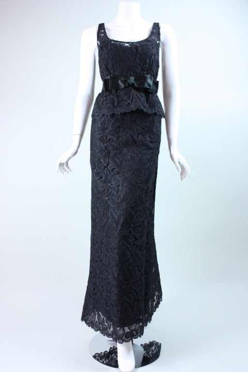 Stunning gown circa 1960's through early 1970's from American designer Mollie Parnis is made out of black raised floral lace.  Fitted bodice is sleeveless, has scoop neck, black satin waistband, and peplum-like detail at the hip.  Straight skirt is
