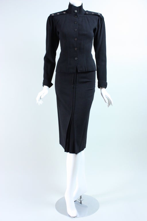 Military-inspired ensemble from Jean Muir dates to the 1970's.  It is made out of black crepe with textured transparent gray buttons.  Fitted jacket is single-breasted, has Mandarin collar, and long sleeves with rolled cuffs.  Slightly flared skirt