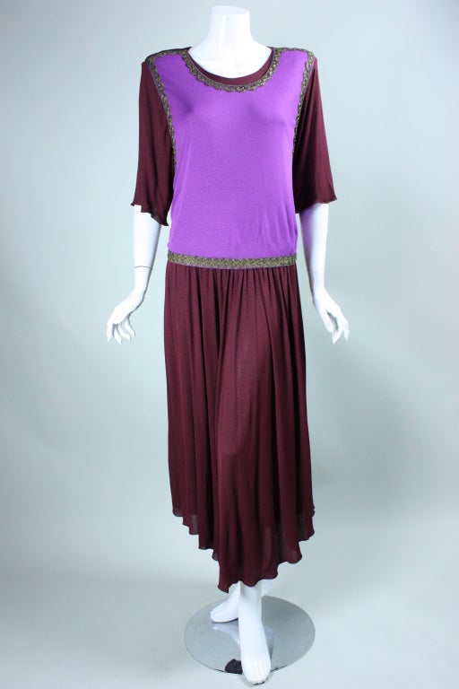 Grecian-influenced dress from Holly's Harp dates to the 1970's.  The dress is made out of burgundy matte jersey with a purple overlayer that is edged with metallic gold trim and ties at the center back waist.  Scoop neck.  Short sleeves.  Padded