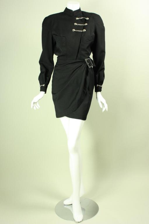Military-inspired dress from Thierry Mugler dates to the 1980's through 1990's.  It is made of black gabardine with silver-toned chain hardware.  Bodice has mandarin collar, long sleeves with button cuffs, two breast patch pockets, and asymmetrical