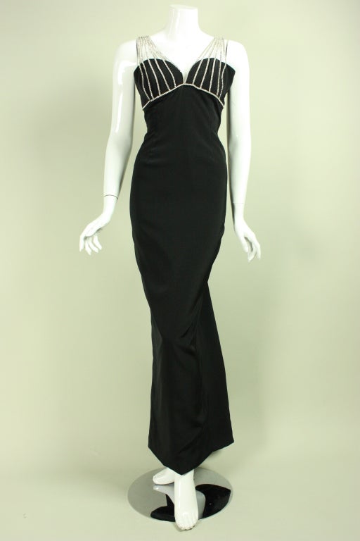 Dramatic gown from Vicky Tiel dates from the 1980's through the 1990's.  It is made of black four-ply silk with a black taffeta godet in the center back.  Bands of clear rhinestones cover the bust like a cage and converge into wide straps.  Center
