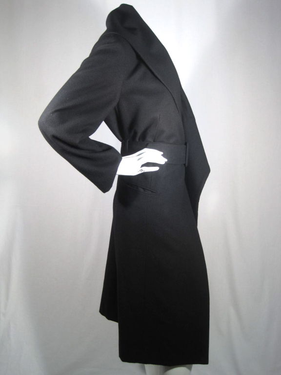 Black asymmetrical belted wool coat from Comme des Garçons has a loosely structured oversized lapel, lightly padded shoulders and front flap pockets. Coat features a belt that cleverly feeds inside through the side seam, cinches the waist and