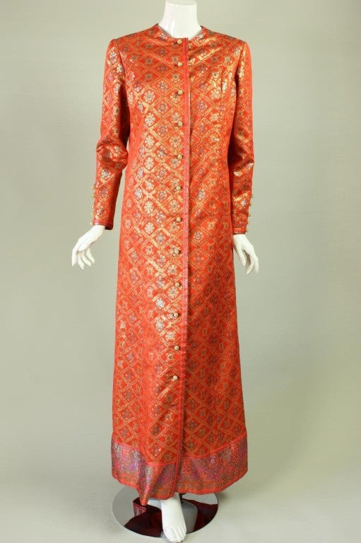 Brightly-colored coat from Oscar De la Renta dates to the 1970's.  It is made of bright orange jacquard with a geometric design in green and purple.  Round neck.  Long, tapered sleeves with buttoned cuffs.  Center front closure with spherical