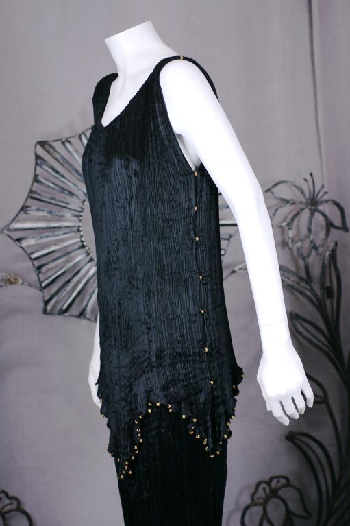 Este vestido está hecho de seda negra finamente plisada con cordones de seda a lo largo de las costuras laterales, los hombros y el dobladillo de la túnica, con cuentas de cristal multicolores enhebradas a través de los cordones. <br />
<br />
El