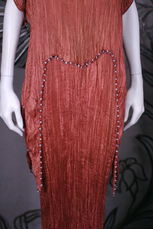 Cette robe est réalisée en soie finement plissée de couleur sienne avec un cordon de soie le long des coutures latérales, des épaules et de l'ourlet de la tunique avec des perles de verre multicolores enfilées dans le cordon.<br />
<br />
Le