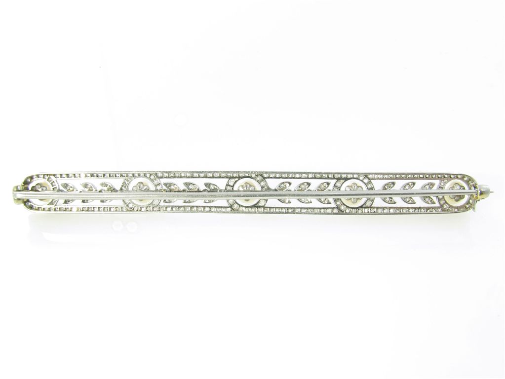 An Edwardian natural pearl, platinum and diamond bar pin.  Circa 1910.  The bar style brooch is set with 5 pearls, each measuring approximately 4.9 mm, set at equal intervals in a rose cut diamond geometric and leaf tip style mount.  The platinum