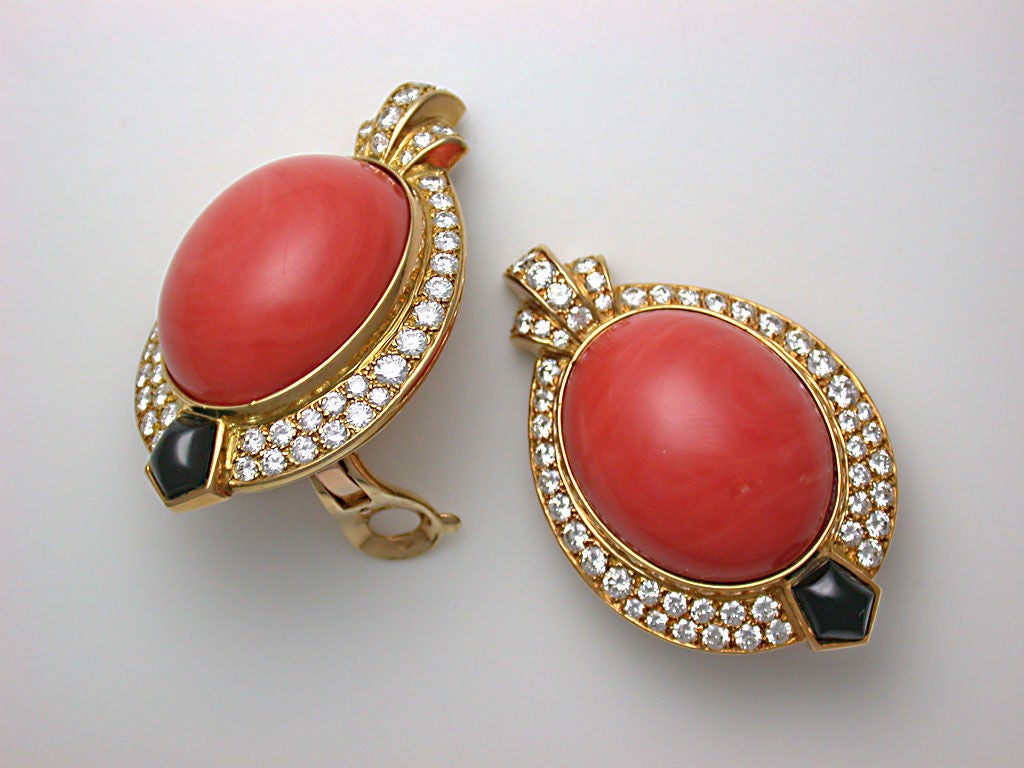 The dramatic, large earclips set to the centers with high-dome medium orange coral cabochons, within oval frames pavé-set with circular-cut white diamonds, further accented with a diamond flourish at the tops and black onyx at the bottoms, signed